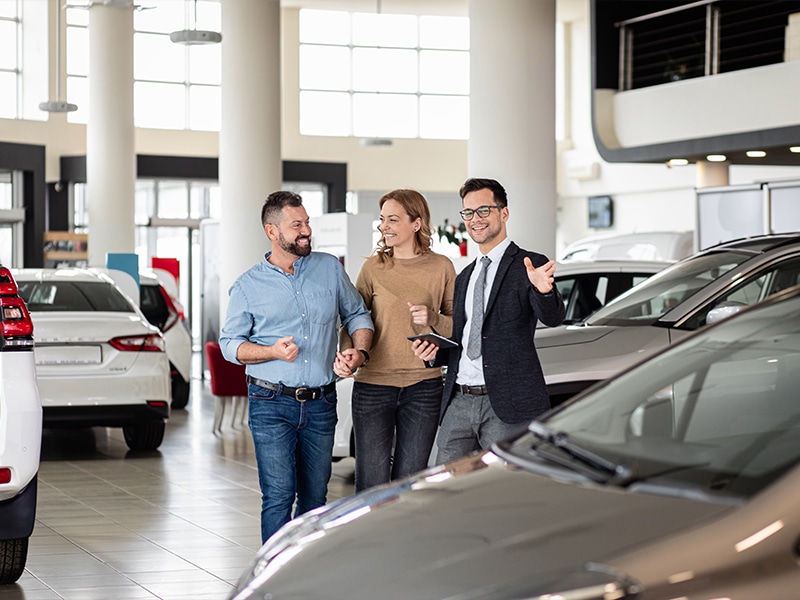 A GMC salesman is showing a smiling couple around the SUVs in the showroom